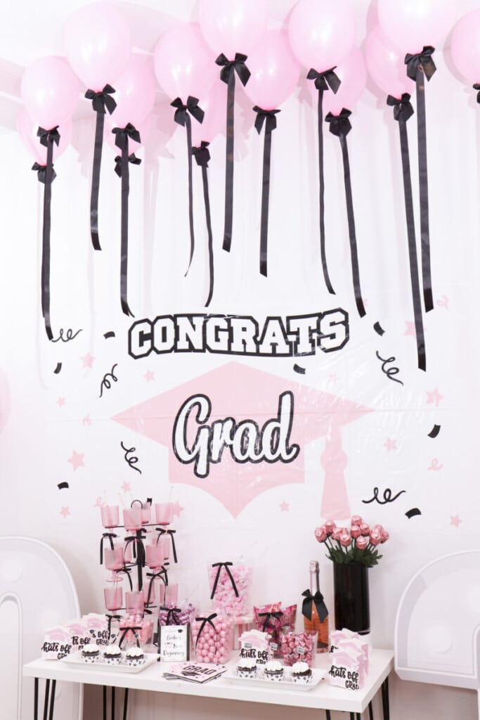 pink and black graduation party candy bar in front of Congrats Grad sign backdrop with ceiling balloons