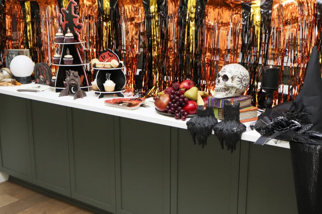 witch party dessert bar on kitchen island with orange, black and gold tinsel backdrop to hide kitchen appliances behind it